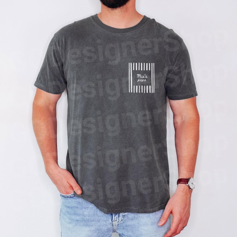 May include: Dark gray t-shirt with a white vertical striped square design on the chest. The square contains the text "Mia's papa" in a handwritten font. The shirt has short sleeves and a crew neck.