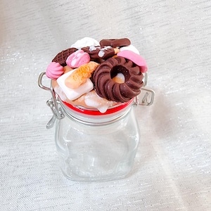 Puede incluir: Un tarro de vidrio con tapa roja y una variedad de galletas de arcilla polimérica hechas a mano y de colores en la parte superior. Las galletas tienen forma de rosquillas, cuadrados y círculos.