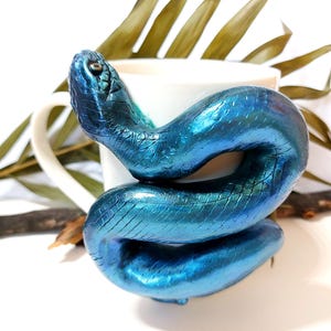 Puede incluir: Una figura de serpiente metálica azul enrollada alrededor de una taza blanca. La serpiente tiene una apariencia texturizada y escamosa y está colocada para que parezca que se envuelve alrededor de la taza. El fondo incluye hojas secas y una superficie blanca.