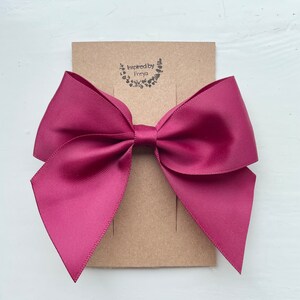 May include: A large burgundy satin bow with a simple design. The bow is attached to a brown card with the text "Inspired by Freya" printed on it.