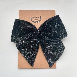 Puede incluir: Un gran lazo de terciopelo negro con purpurina plateada. El lazo está sobre una tarjeta marrón con el texto "inspired by Freya" impreso en ella.
