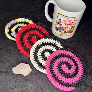 Puede incluir: Cinco posavasos de ganchillo con un diseño en espiral blanco y negro. Los posavasos son de diferentes colores: rosa, rojo, blanco y azul claro. Los posavasos están dispuestos en círculo sobre una superficie negra.