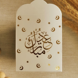 May include: White gift bag with a gold foil design of crescent moons and stars. The bag has the Arabic text "Eid Mubarak" in gold foil.