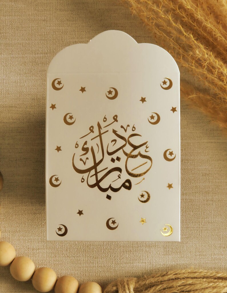 May include: White gift bag with a gold foil design of crescent moons and stars. The bag has the Arabic text "Eid Mubarak" in gold foil.