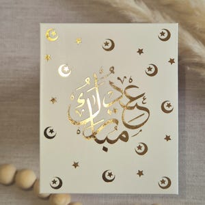 May include: A white box with gold foil lettering that says "Eid Mubarak" in Arabic script. The box is decorated with gold foil stars and crescent moons.