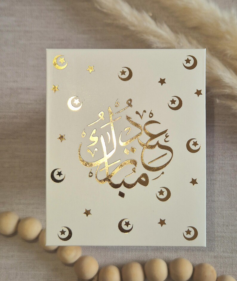May include: A white box with gold foil lettering that says "Eid Mubarak" in Arabic script. The box is decorated with gold foil stars and crescent moons.