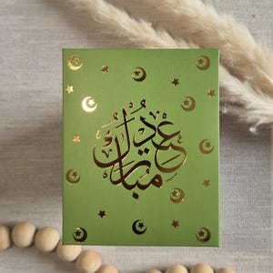 May include: A green card with gold foil lettering that says "Eid Mubarak" in Arabic script. The card is decorated with gold foil stars and crescent moons.