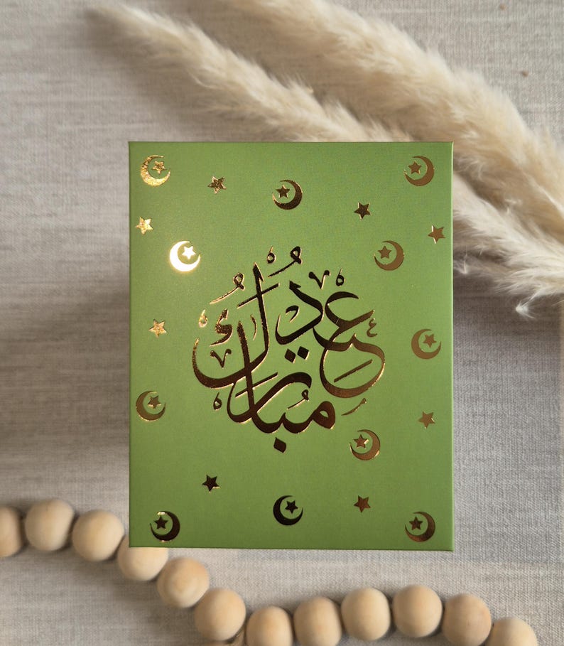 May include: A green card with gold foil lettering that says "Eid Mubarak" in Arabic script. The card is decorated with gold foil stars and crescent moons.
