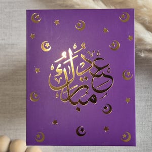 May include: Purple card with gold foil accents. The card features a crescent moon and star design and the Arabic text "Eid Mubarak" in gold foil.