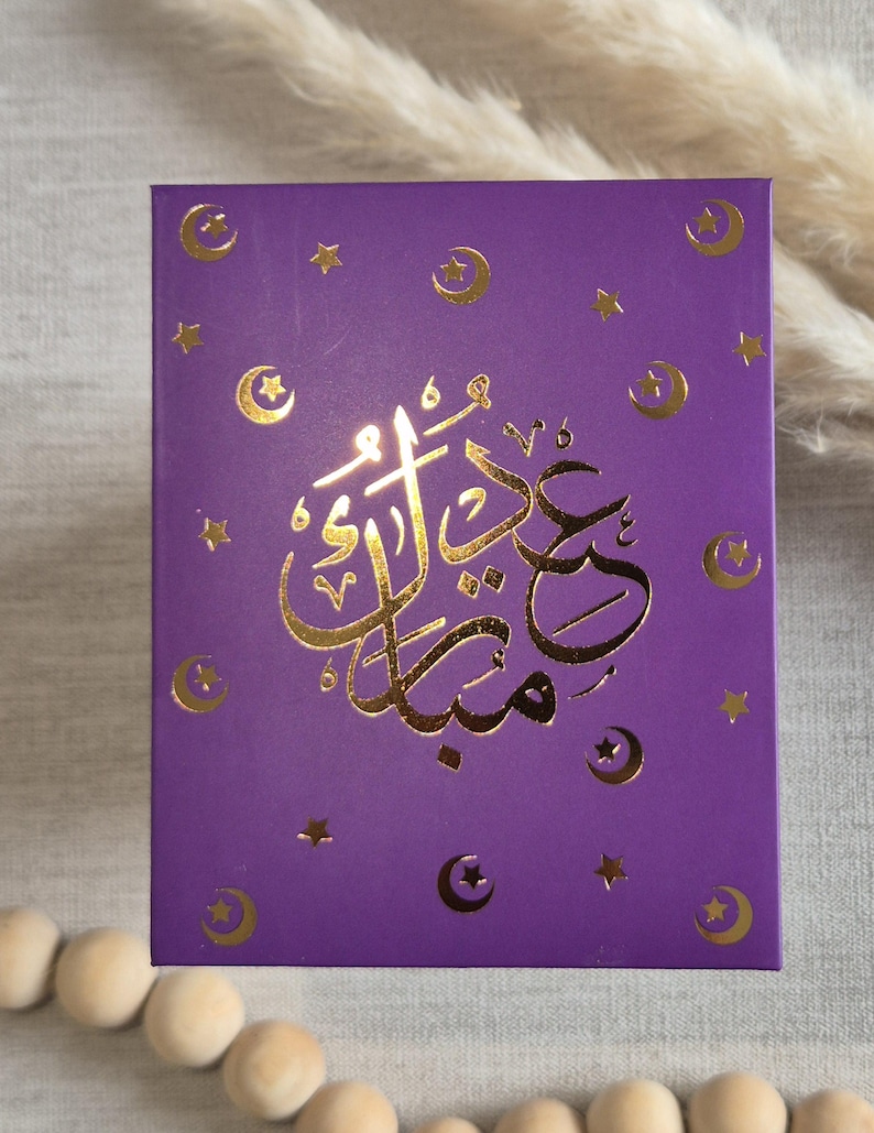 May include: Purple card with gold foil accents. The card features a crescent moon and star design and the Arabic text "Eid Mubarak" in gold foil.
