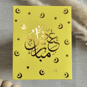 May include: A yellow card with gold foil stars and crescent moons. The card has the Arabic text "Eid Mubarak" in gold foil.