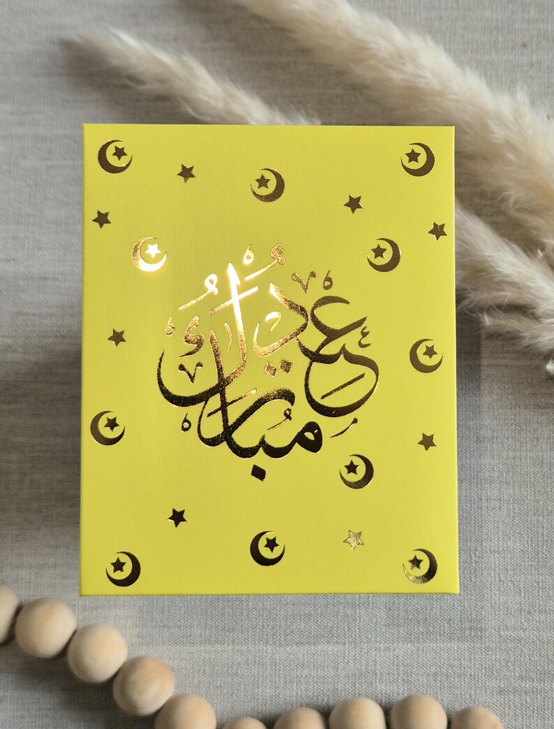 May include: A yellow card with gold foil stars and crescent moons. The card has the Arabic text "Eid Mubarak" in gold foil.