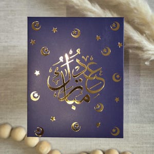 May include: A dark blue card with gold foil lettering and crescent moon and star designs. The card says "Eid Mubarak" in Arabic.