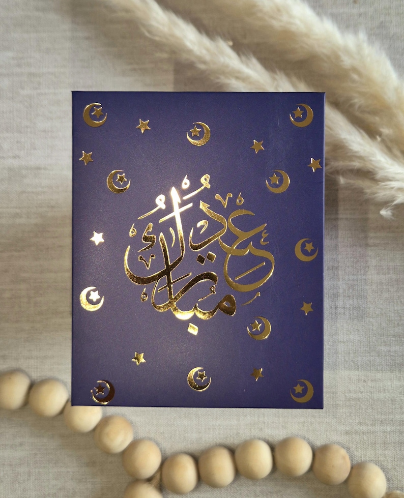 May include: A dark blue card with gold foil lettering and crescent moon and star designs. The card says "Eid Mubarak" in Arabic.