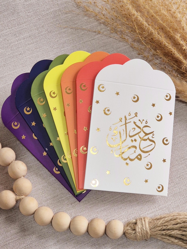 May include: Set of eight colourful envelopes with gold stars and crescent moons. The envelopes are folded in half and have a scalloped edge. The top envelope has the Arabic text "Eid Mubarak" in gold foil.
