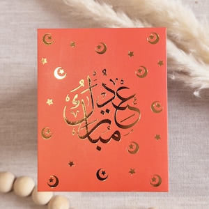 May include: A red card with gold foil lettering and stars. The card says "Eid Mubarak" in Arabic.