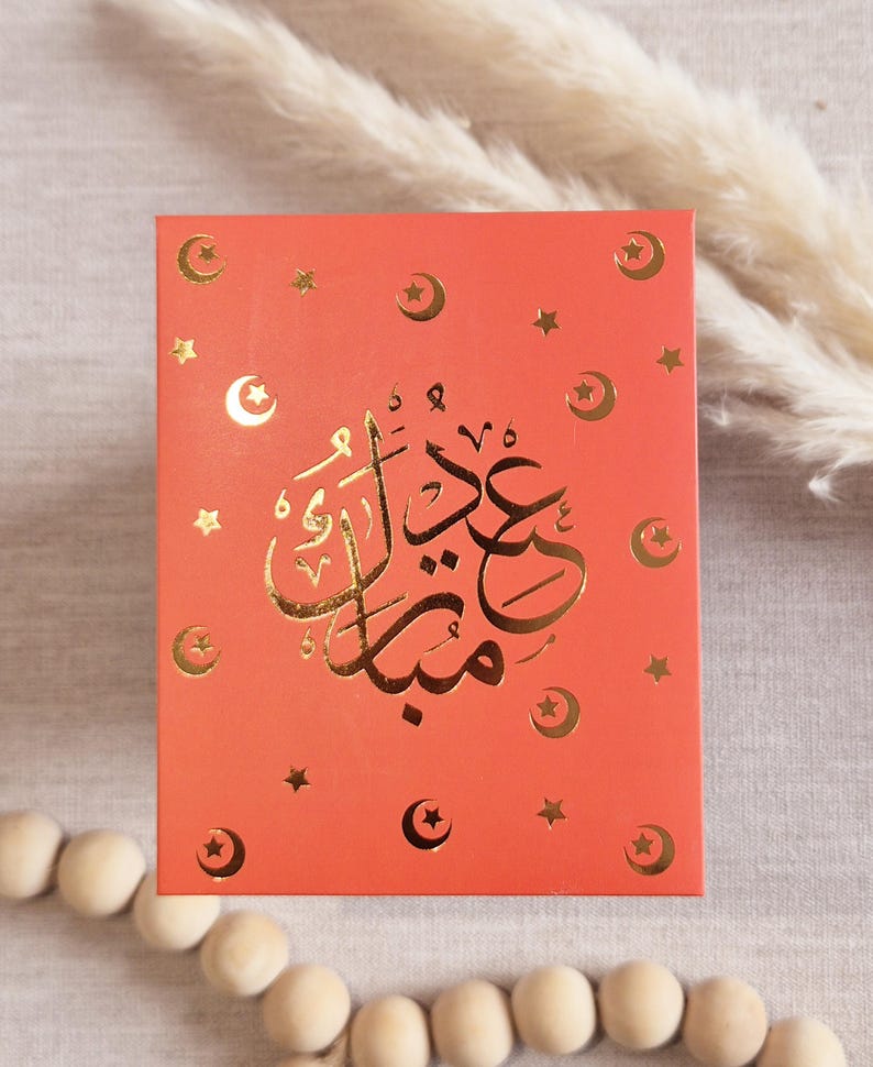 May include: A red card with gold foil lettering and stars. The card says "Eid Mubarak" in Arabic.