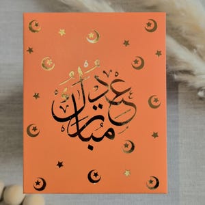 May include: An orange card with gold foil accents. The card features a crescent moon and star design and the Arabic text "Eid Mubarak".