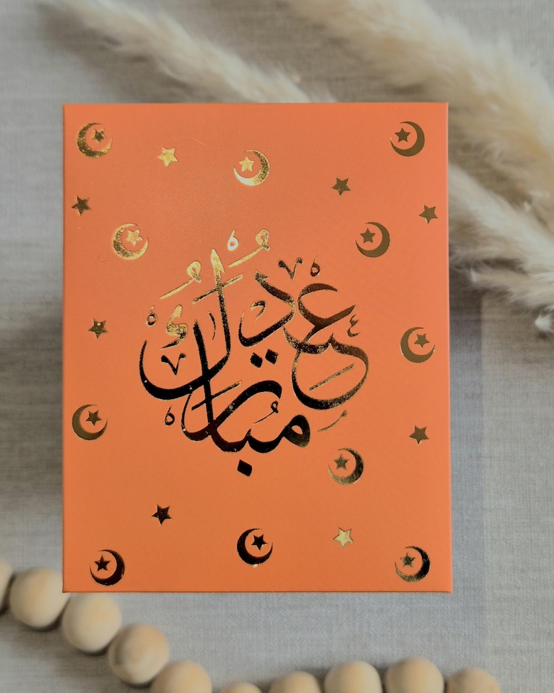 May include: An orange card with gold foil accents. The card features a crescent moon and star design and the Arabic text "Eid Mubarak".