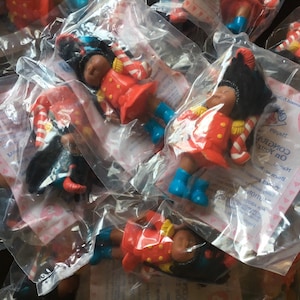 May include: A pile of plastic bags containing small, red and blue toy figures of a girl wearing a red and white uniform with a candy cane. The bags have a pink and white background with the text "Happy Holidays On Your Way!" and a heart with the letter "C" inside.