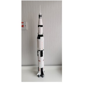 May include: A white and black model of the Saturn V rocket with the words "United States" and "USA" printed on the side. The rocket has a pointed nose cone and four engines.