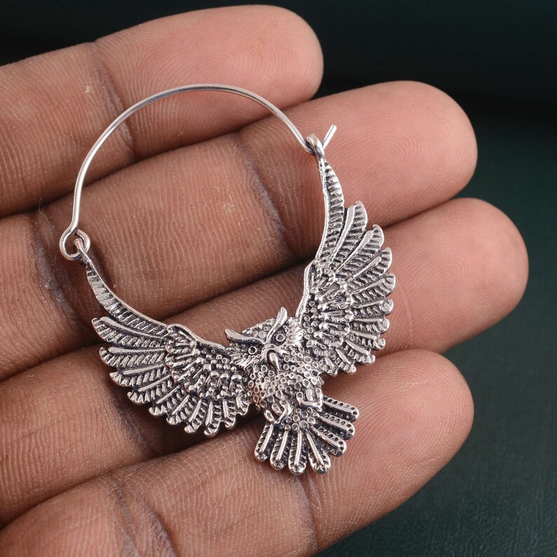 May include: Silver hoop earrings with an owl design. The owl is in flight with its wings spread wide. The earrings are made of metal and have a silver finish.