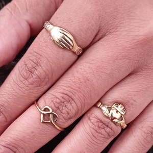 May include: Three gold rings on a hand. The rings are a heart-shaped ring, a ring with a clasped hands design, and a Claddagh ring with a heart and two hands.