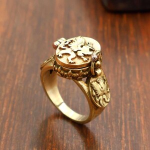 May include: A gold-colored ring with a detailed design. The ring features a lion emblem on the top and decorative elements on the band. The ring appears to be made of metal and has an antique or vintage aesthetic.