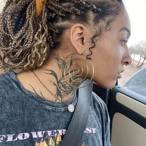 May include: A person with long, braided hair styled in a bun, wearing a grey t-shirt with a sunflower graphic and the text "FLOWER", and a gold spiral hoop earring.
