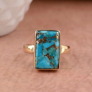 May include: A gold ring featuring a rectangular turquoise gemstone. The turquoise has a marbled pattern with gold flecks.