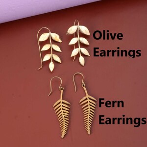 Puede incluir: Dos pares de pendientes de color dorado. El primer par presenta un delicado diseño de rama de olivo. El segundo par presenta un diseño de hoja de helecho. El texto "Olive Earrings" y "Fern Earrings" es visible en la imagen.