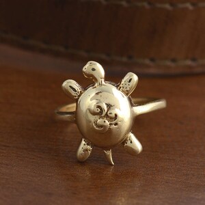 May include: A gold-toned turtle ring with a detailed design on the shell. The ring is made of metal and has a simple band.