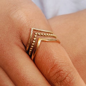 May include: Three gold rings stacked on a finger. The top ring is a thin, open triangle shape. The middle ring is a band of small, round beads. The bottom ring is a thicker, solid band.