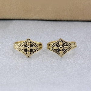 Puede incluir: Dos anillos de dedo del pie de oro con un diseño en forma de diamante que presenta una flor en el centro.