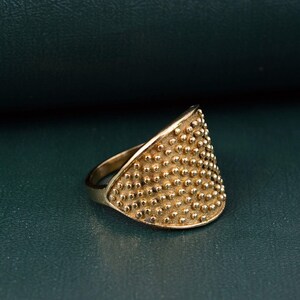 May include: A gold-toned ring featuring a wide, curved front panel with a textured surface covered in numerous small, raised spherical studs. The band of the ring is smooth and tapers toward the back, creating a distinct contrast with the detailed front.