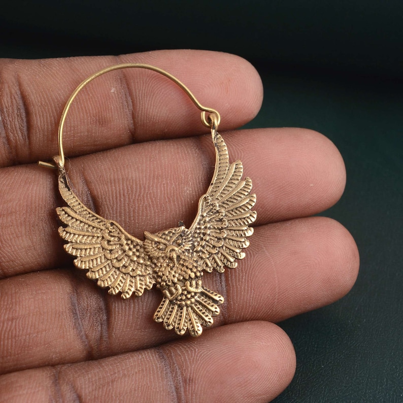 May include: Gold hoop earrings with an owl design. The owl is detailed with feathers and has its wings spread out.