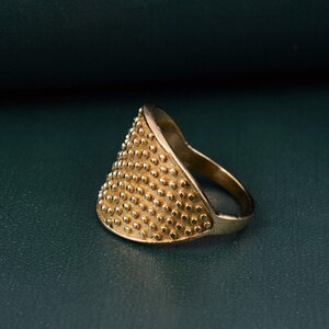 May include: A gold-toned ring featuring a wide, triangular-shaped face covered in a dense, uniform pattern of small, raised circular studs. The band of the ring is smooth and tapers toward the back, creating a distinct contrast with the textured front.