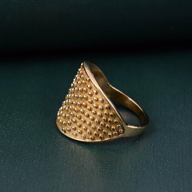 May include: A gold-toned ring featuring a wide, triangular-shaped face covered in a dense, uniform pattern of small, raised circular studs. The band of the ring is smooth and tapers toward the back, creating a distinct contrast with the textured front.
