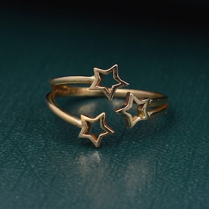 Könnte beinhalten: Ein goldfarbener Ring mit drei Sternaussparungen. Der Ring hat ein Wickeldesign mit zwei Bändern. Die Sterne sind gleichmäßig um den Ring herum angeordnet. Der Ring befindet sich vor einem dunkelblauen Hintergrund.