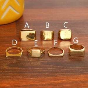 May include: Seven gold-coloured rings in various designs are displayed. Styles include rectangular, square, and rounded shapes. The rings are labelled A to G. They are presented on a wooden surface.