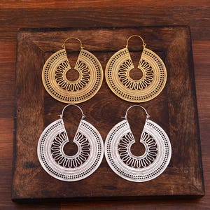 May include: Two pairs of large circular hoop earrings, one pair in a gold tone and one pair in a silver tone. Each earring features a central circular cutout surrounded by a wavy line pattern, enclosed by a wide, intricate lattice-work border.