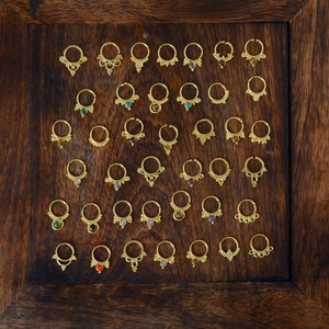 Puede incluir: Una muestra de anillos de nariz de oro con diversos acentos de piedras preciosas. Los anillos están dispuestos en filas sobre un fondo de madera.