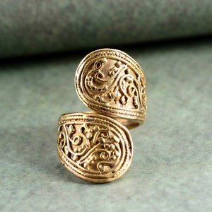 May include: A gold-colored ring with an ornate, swirling floral design. The ring has an open, adjustable design. The metal has a slightly aged appearance, with detailed patterns throughout the surface.