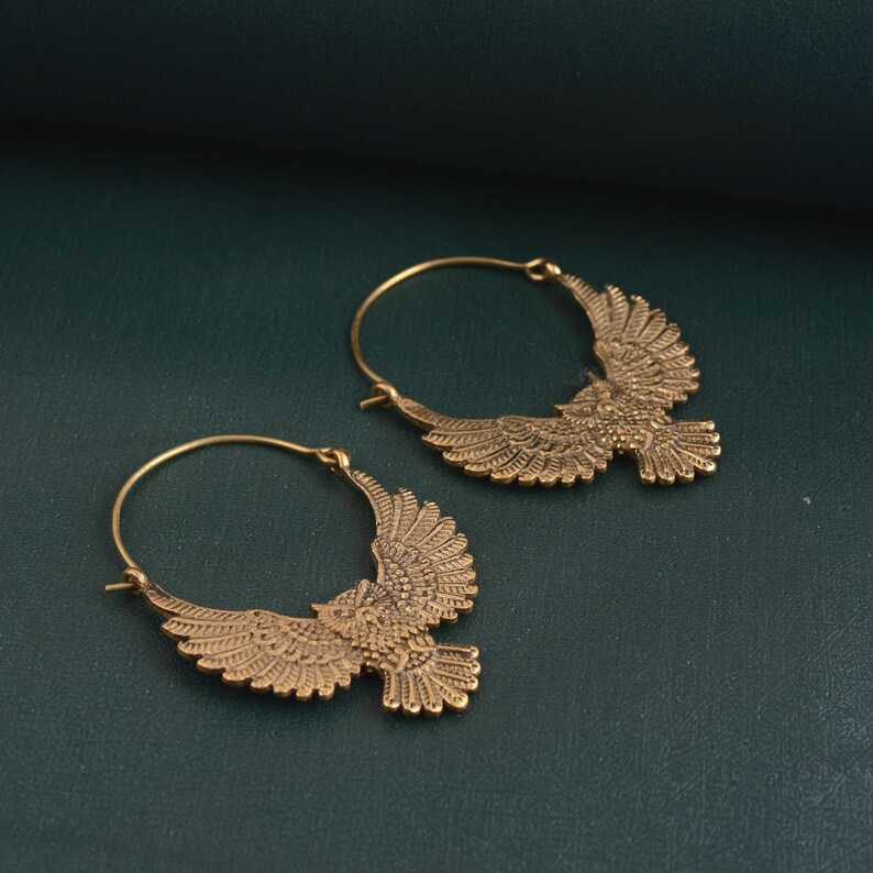 May include: A pair of gold hoop earrings with a detailed eagle design. The eagle is in flight with its wings spread wide.