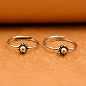 May include: Two adjustable toe rings with a sunflower design. The rings are made of a gold-colored metal and feature a small, round, gold-colored center surrounded by detailed petals. The rings are displayed on a brown surface.
