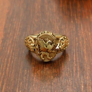 May include: A gold-colored ring with an oval centerpiece featuring a detailed griffin design. The ring has ornate side detailing and a slightly worn appearance, suggesting an antique or vintage style. The ring is set against a dark wood surface.
