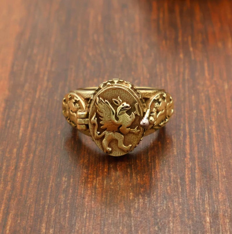 May include: A gold-colored ring with an oval centerpiece featuring a detailed griffin design. The ring has ornate side detailing and a slightly worn appearance, suggesting an antique or vintage style. The ring is set against a dark wood surface.