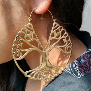 Puede incluir: Pendientes de aro dorados con un diseño de árbol de la vida. Los pendientes tienen un patrón calado detallado con forma circular. El diseño del árbol está centrado dentro del aro, con ramas y raíces. Los pendientes se llevan en la oreja.