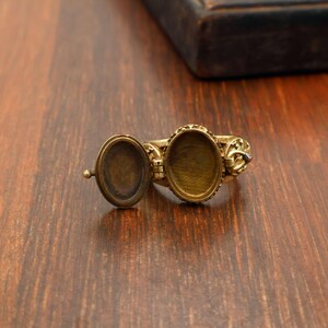 May include: A vintage-style brass locket ring with an oval compartment that opens. The ring has an ornate design around the oval frame and on the band. The ring is open, revealing the interior of the locket.