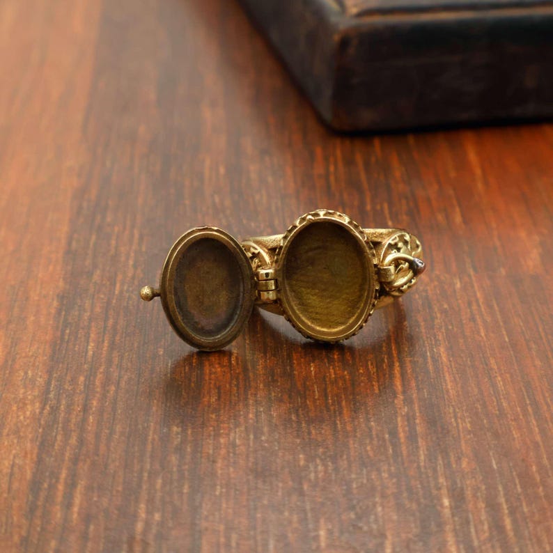 May include: A vintage-style brass locket ring with an oval compartment that opens. The ring has an ornate design around the oval frame and on the band. The ring is open, revealing the interior of the locket.